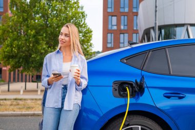 Sarışın kadın şarj olan elektrikli bir arabanın yanında duruyor, kahvesini yudumluyor ve sokakta akıllı telefonunu kontrol ediyor..