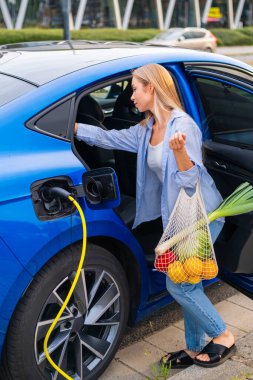 Kadın arka koltuktan taze yiyecek indirirken elektrikli aracını fişe takıyor.
