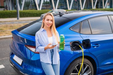 Bir kadın elektrikli arabasının yanında duruyor, elinde yeşil bir şişeyle şarj oluyor ve parasını ödemek için telefonunu kullanıyor. Yeşil enerji kavramı