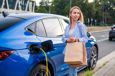 Kadın elektrikli arabasını park eder ve akşam alışveriş torbaları taşırken şarj etmeye başlar.