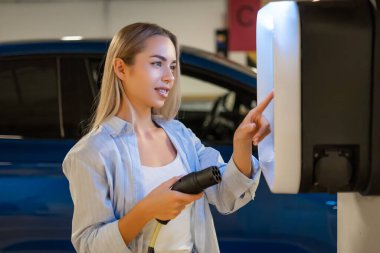 Kadın bir otoparkta elektrikli aracı için şarj istasyonu kullanıyor.