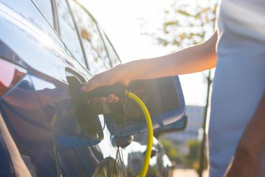 Kadın eli, güneş ışığına karşı elektrikli arabayı şarj etmek için kabloyu bağlar.. 