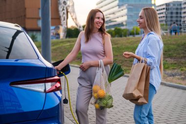 Biri otoparkta elektrikli bir arabayı şarj ederken, iki kadın muhabbet eder ve ellerinde alışveriş torbalarıyla dolaşır.