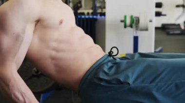 Eşofmanlı bir adam spor salonunda bacak barfiks çekerek mekik çekiyor. Çıplak gövdeye sahip sporcu karın kaslarını güçlendirir. Göğüs ve bacaklarla hareket eder.