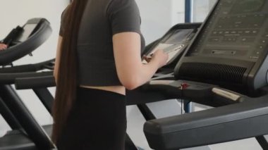 Akıllı telefonlu bir bayan sporcu spor salonunda büyük bir koşu bandında yürüyor. Bayan boş zamanlarını spor yaparak geçiriyor bacak kaslarını ve ses tonunu güçlendiriyor.