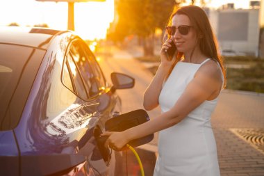 Sokakta gün batımında cep telefonuyla konuşan bir kadın elektrikli arabayı şarj ediyor.