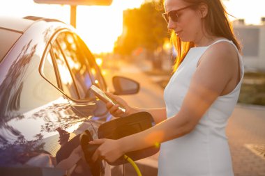 Beyaz elbiseli kadın gün batımında elektrikli arabasını şarj ederken cep telefonu kullanıyor.