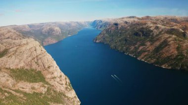 Görkemli Lysefjord berrak mavi gökyüzünün altında engebeli dağları incelikle kesiyor. Uzak teknelerden uyanan fiyordun derin mavi suları