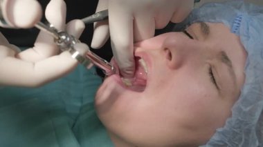 Diş hekimi hasta diş etlerine lokal anestezi uygular. Ameliyat hazırlıkları sırasında ağzı açık, mavi cerrahi şapkalı bir kadın.