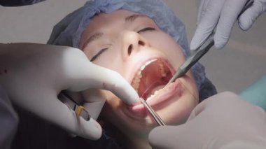 Dişçi, penslerle ince iplikleri manipüle eden bir kadın üzerinde çalışıyor. Asistan emme aracı ile alanı yönetmeye yardımcı olur ve net görünürlüğü korur