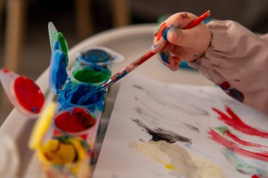 Gouache renginde bir boya fırçasına uzanan küçük bir çocuğun eli. Evde yaratıcı aktivite, erken çocukluk sanatı eğitimi.