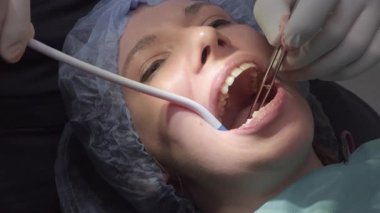 Dentist works in patient mouth using suction tool and surgical tools. Woman lies in dental chair in surgical cap with mouth open during procedure