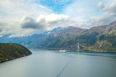 Distant tourist ship sails under Hardanger Bridge gracefully spanning waters of fjord. Mountains with snow on tops create Norwegian landscape