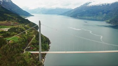 Hardanger Köprüsü, kıyı şeridinden yükselen yüksek kulelerle geniş fiyort boyunca uzanır. Köprü, yeşil kara parçalarını karla kaplı dağlara bağlıyor.