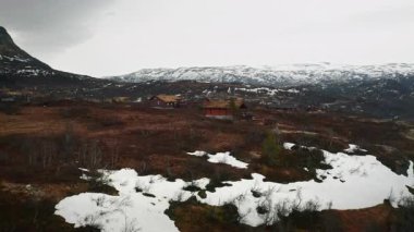 Dağlık vadide otlak çatıları olan geleneksel Norveç evleri. Yerdeki kar parçaları bulutlu gökyüzünün altındaki uzak tepelere kadar uzanır.