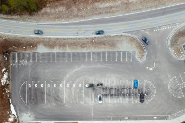 Elektrikli araç park alanındaki şarj istasyonuna yaklaşıyor. Birçok park çizgisi olan gri asfalt modern tesiste sessiz bir an olduğunu gösteriyor.