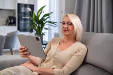 Gülümseyen orta yaşlı kadın tabletinde film seyrediyor, evdeki rahat bir koltukta oturuyor. Eğlence, eğlence ve modern teknoloji konsepti.