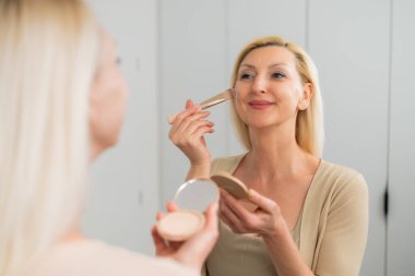  Açık renk elbiseli güzel bir kadın, pürüzsüz cilt için makyaj fırçası kullanıyor.