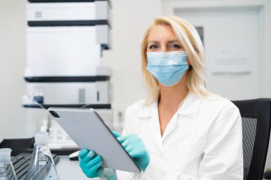 Blonde female scientist wearing a lab mask and rubber gloves recording chemical experiment results on a tablet while standing in a chemistry laboratory with instruments around.