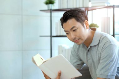 Mutlu Asyalı genç adam kütüphanede ya da dinlenme odasında ders kitabı okuyor..
