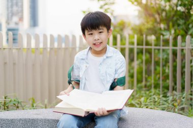Neşeli mutlu Asyalı çocuk bankta oturuyor ve halk parkında tek başına kitap ya da masal okuyor..