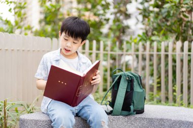Neşeli mutlu Asyalı çocuk bankta oturuyor ve halk parkında tek başına kitap ya da masal okuyor..