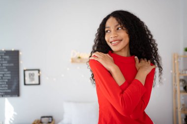 Mutlu - neşeli, güzel siyah kız Noel temalı bir yatak odasında kırmızı bir kazak giyiyor, güzel bir kız yatak odasında kollarını kavuşturuyor ve kameraya güzel bir gülümseme yapıyor..