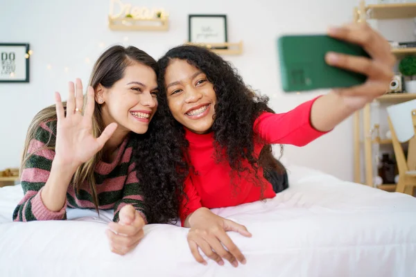 Happy diverse women black and caucasian laying down on the bed and making a video call on smartphone with a friends in Christmas and New Year festival. Diverse ethnics women talking together.