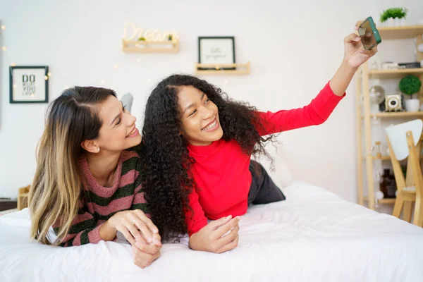 Happy diversity women black and caucasian laying down on the bed and making a video call on smartphone with a friends in Christmas and New Year festival. Diverse ethnics women talking together.