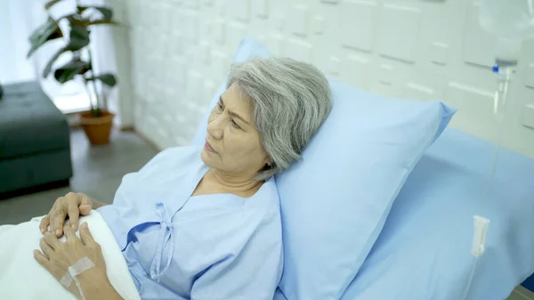 Unhappy - lonely Asian senior elder woman patient looking outside the window during resting in the recovery ward in hospital. Lonely elder woman feels sad and emotional. Mental health concept.
