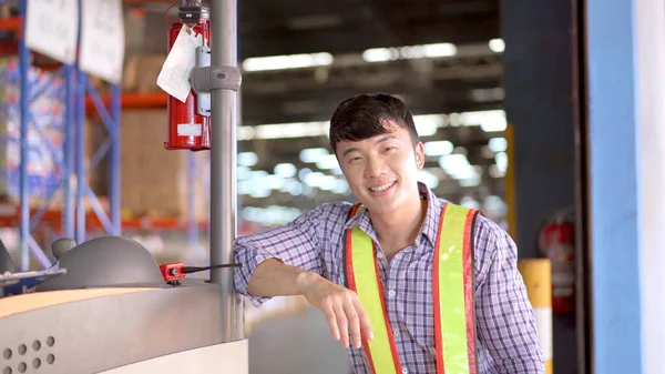 Happy and satisfied Asian worker or staff in warehouse standing beside a forklift cart and smiling to camera for portrait, photograph with copyspace. Skillful Asian working in a factory driving a forklift.