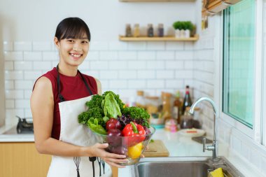 Yemek pişirmeden önce sağlıklı bir Asyalı kadın mutfak musluğunda sebze yıkıyor. Neşeli, sağlıklı Asyalı kadın bir sepet sebze ve meyve taşıyor ve kameraya gülümsüyor..