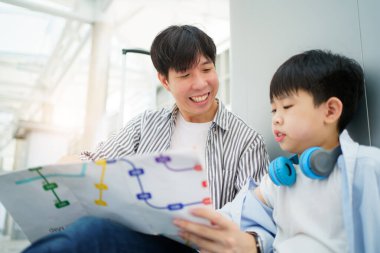 Mutlu Asyalı aile şehir merkezinde birlikte seyahat ediyor. Baba, oğluyla birlikte tren istasyonunun haritasına bakıyor ve haritada işaret parmağıyla işaret ediyor. Şehirdeki Asyalı aile turisti.