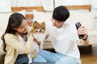 Mutlu Asyalı genç çift selfie çekerken ya da akıllı telefondan köpekleriyle oturma odasında selfie çekerken. Kadın ve erkek akıllı telefonda kameraya gülümsüyor..