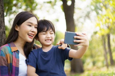 Neşeli Asyalı küçük çocuk, sabahleyin annesiyle parkta fotoğraf çektirmek için akıllı telefon kullanıyor. Mutlu aile zamanı konsepti, hafta sonu küçük oğluyla kalan bir kadın..