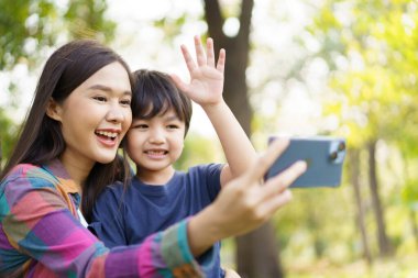 Neşeli Asyalı küçük çocuk, sabahleyin annesiyle parkta fotoğraf çektirmek için akıllı telefon kullanıyor. Mutlu aile zamanı konsepti, hafta sonu küçük oğluyla kalan bir kadın..