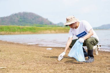 Asyalı genç adam plajda plastik şişe çöpü topluyor, erkek gönüllü sahilde yürüyor ve plastik atık topluyor..