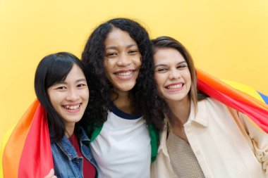 Mutlu mutlu LGBTQ çok ırklı kadınlar fotoğraf için poz veriyor ve sarı arka planda gökkuşağı bayrağı var. LGBTQ insanlarının ifade özgürlüğü.