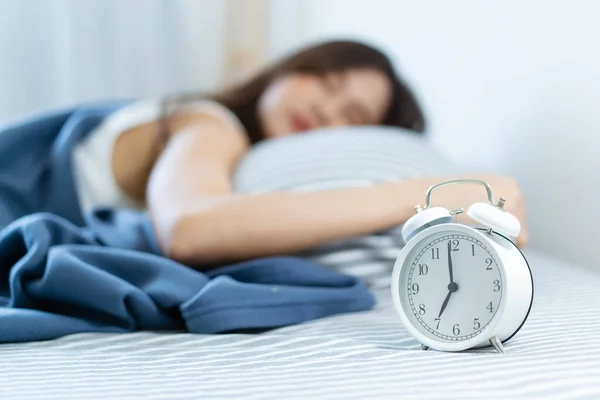 Asian young woman sleeping on the cozy bed in the early morning and trying to turn off or snooze an alarm clock. Lazy sleepy woman wake up late in the morning.