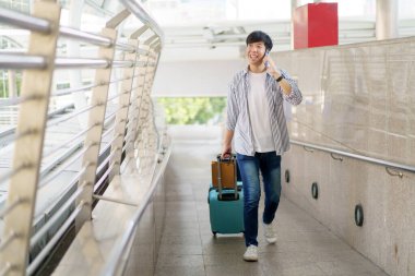 Asyalı genç bir adam valizini şehir boyunca ya da havaalanı terminali boyunca alışveriş torbalarıyla sürüklüyor. Şehirde ya da şehir merkezinde seyahat eden bir adam.