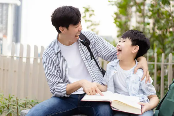 Cheerful happy Asian little boy and his father reading a book or fairy tale together at the outdoor park. Asian man and his son reading a book together.