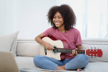 Mutlu Amerikalı Afrikalı siyahi kadın oturma odasında kanepede oturuyor ve akustik gitar çalıyor..
