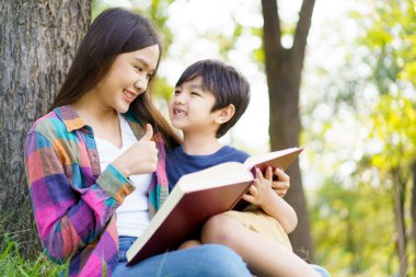 Mutlu Asyalı sevimli küçük çocuk ve anne sabah parkta ağacın altında oturup birlikte kitap okuyorlar. Asyalı kadın hafta sonu sevgili oğluyla vakit geçiriyor..