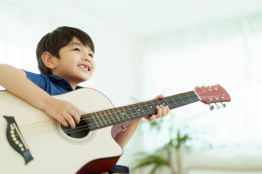 Mutlu Asyalı küçük çocuk yatak odasında akustik gitar çalma alıştırması yapıyor. Gitar çalan sevimli Asyalı çocuk..