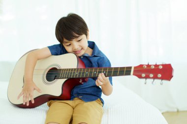 Mutlu Asyalı küçük çocuk yatak odasında akustik gitar çalma alıştırması yapıyor. Gitar çalan sevimli Asyalı çocuk..