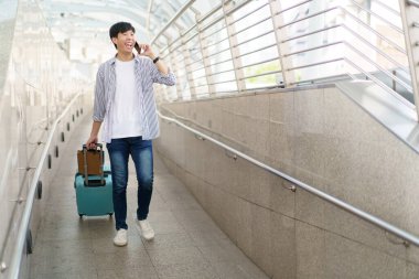 Asyalı genç bir adam valizini şehir boyunca ya da havaalanı terminali boyunca alışveriş torbalarıyla sürüklüyor. Şehirde ya da şehir merkezinde seyahat eden bir adam.