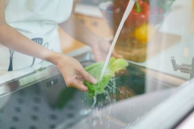 Yemek pişirmeden önce sağlıklı bir Asyalı kadın mutfak musluğunda sebze yıkıyor. Bir ev hanımı sağlıklı bir yemek pişirmek için meyve ve sebzeleri dikkatlice hazırlar ve temizler..