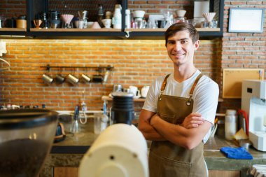 Kahve dükkanındaki tezgahın içinde duran ve kollarını kavuşturup kameraya bakan genç profesyonel kahve barmeni yakışıklı, beyaz tenli barista pozu veriyor..