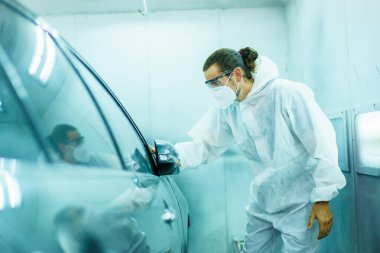 Kimyasal koruma giysisi içindeki profesyonel otomotiv boyama teknisyeni boya odasındaki boya kalitesini inceliyor. Araba boyama ve teknisyenlerin portresi.