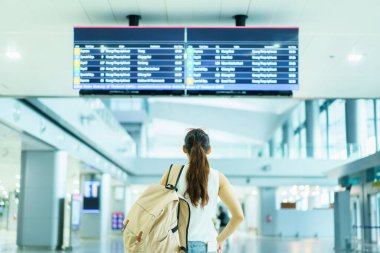 Mutlu Asyalı güzel bayan sırt çantalı turist havaalanı terminalinden kalkış ve varış bilgilerine bakıyor, yalnız kadın turist konsepti. Uçuş programını kontrol eden kadın.